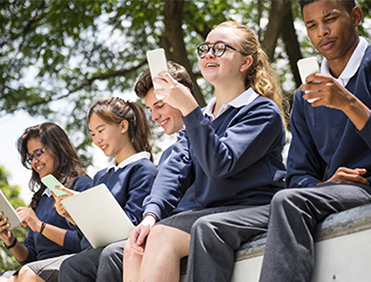 Group of five school friends enjoying their mobile phones and laptops outdoors.