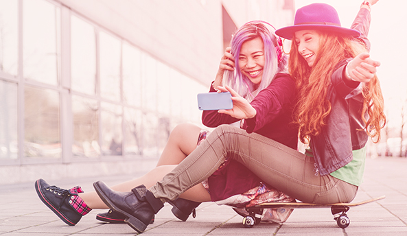Two young women taking photos on their phones whilst sitting on their skateboards.