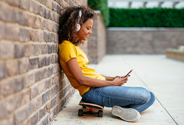 Young girl happily sitting on her skateboard whilst listening to music on her phone.