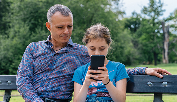Cyberbullying_families_Thumbnail-593x343 A young woman is looking at her smartphone whilst her father pensively looks on.